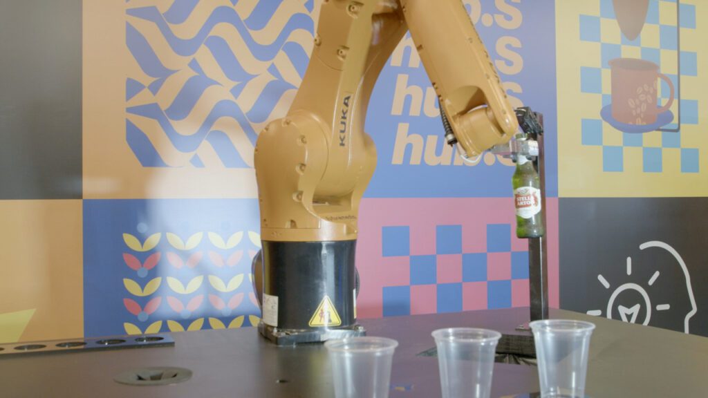 Fotografia colorida do robô que serve cerveja. É um braço mecânico de cor amarela. Na imagem, ele abre uma garrafa. 