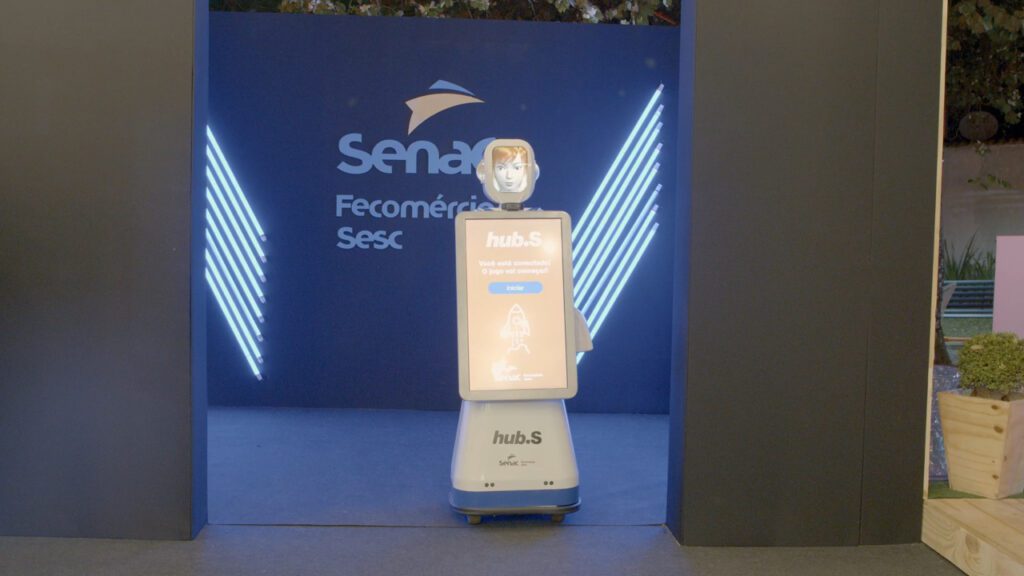Fotografia colorida da robô display, que possui duas telas articuladas, uma maior centralizada e outra acima, simulando a cabeça da robô. Atrás da robô, há a marca do Senac, Fecomércio e Sesc em segundo plano.