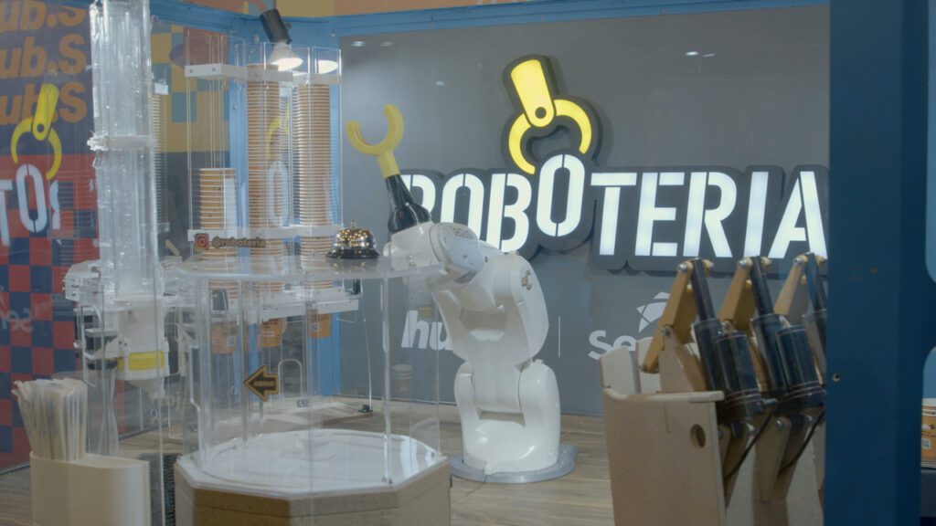 Fotografia colorida da Roboteria, que consiste em um braço mecânico na cor branca e garra amarela. No fundo, há a marca do robô estampada na parede do evento. 