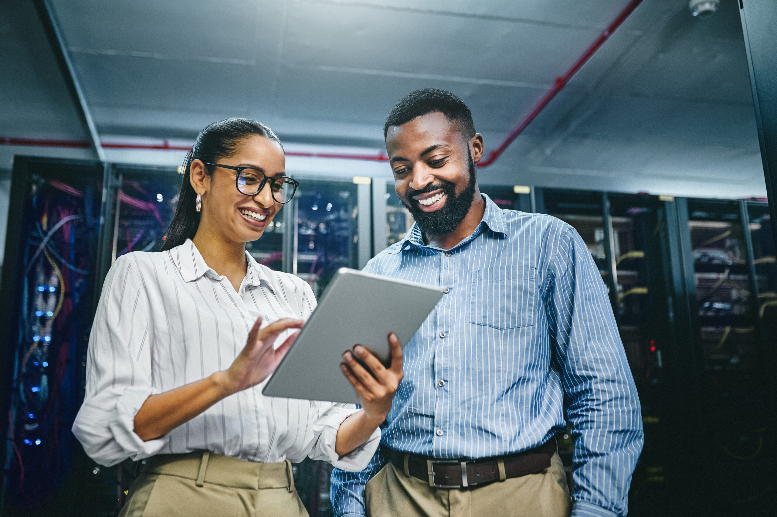 Fotografia colorida em que aparecem um homem e uma mulher. Eles são negros, vestes roupas sociais e olham para a tela de um tablet sorridentes num ambiente corporativo.