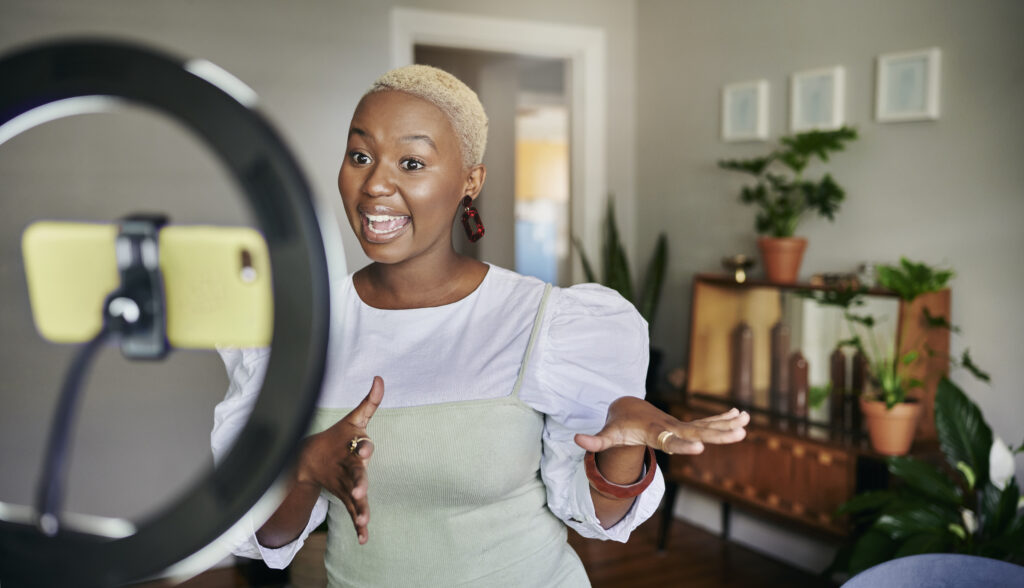 Sorridente jovem influenciadora negra em pé em sua sala de estar em casa e conversando durante uma postagem de vlog usando um smartphone.