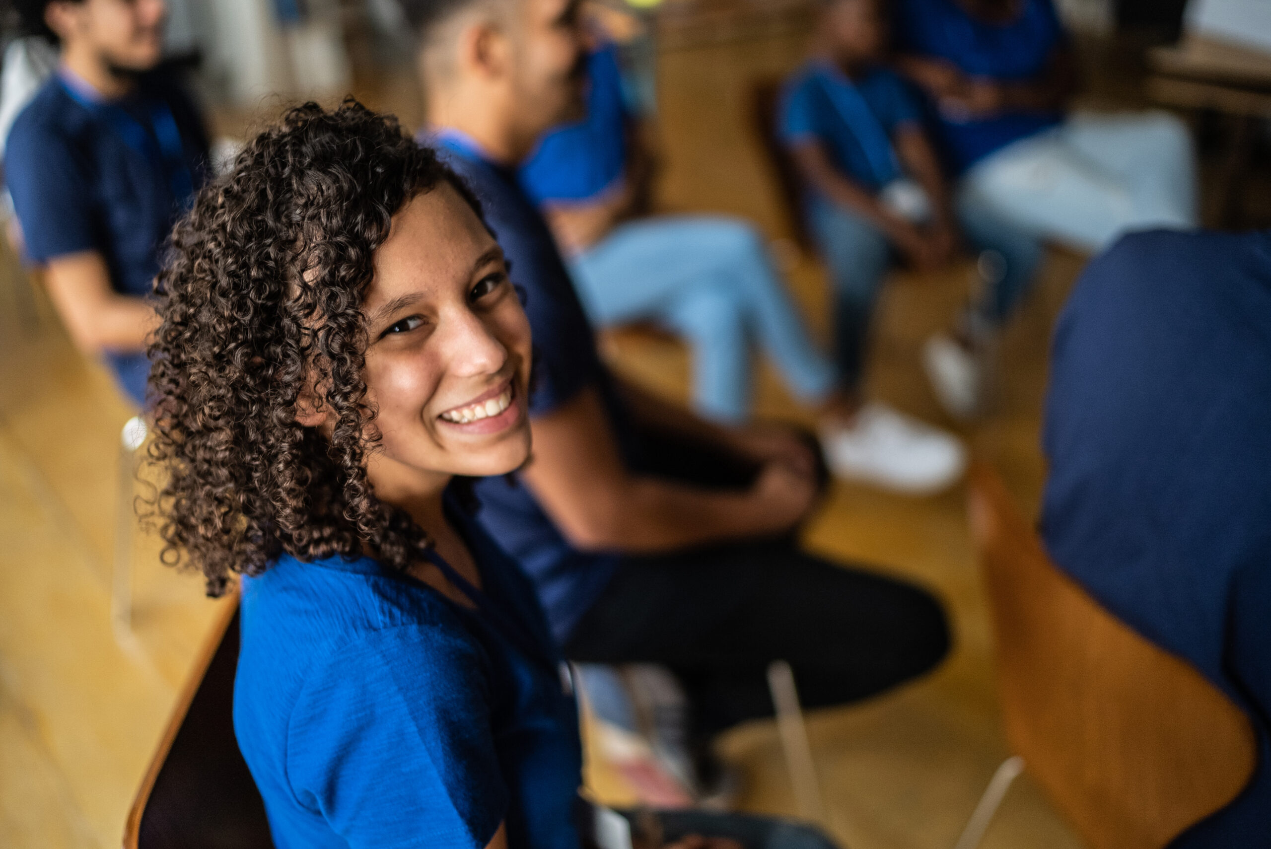 Foto colorida de uma adolescente em uma reunião escolar. Ela é negra, possui os cabelos cacheados, usa uniforme azul e sorri.