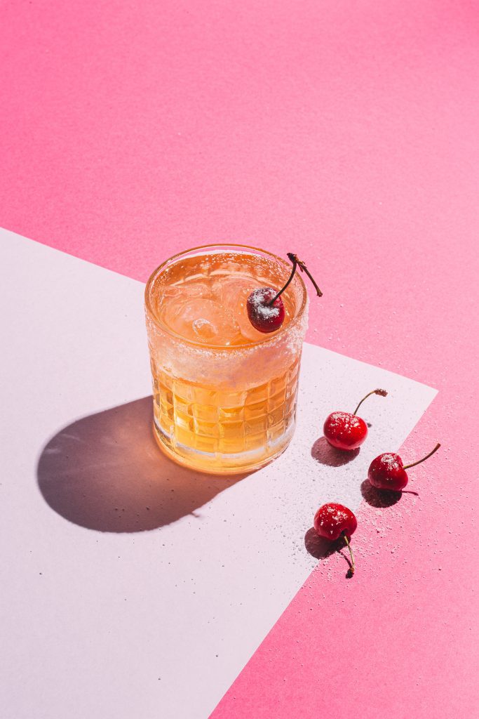 Drink "B.Sour" de cor amarela, com cachaça e decoração com cerejas. Fundo rosa. 